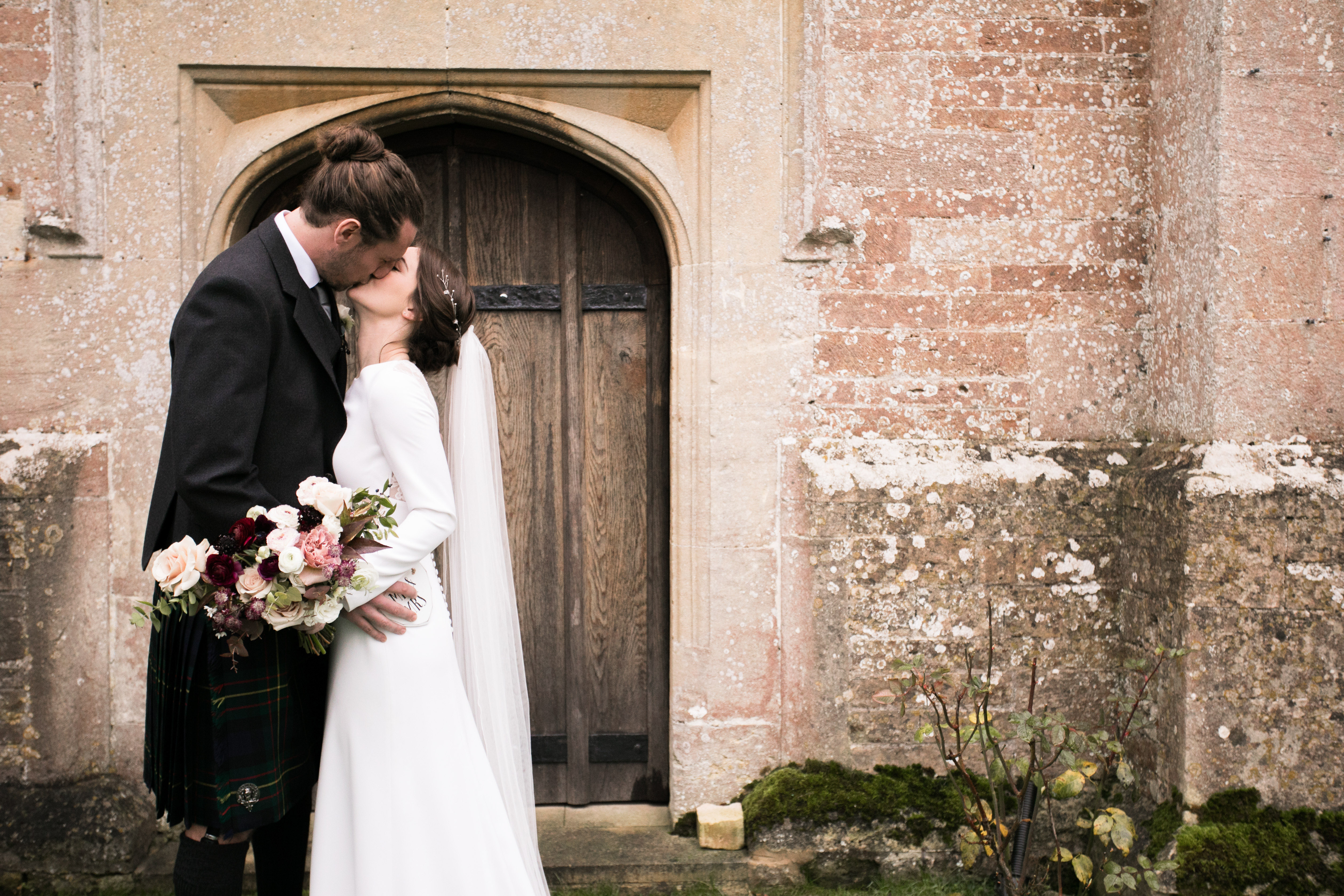 bride and groom kissing in front of an old stone arch with a wintery colour pallette of maroon white and black